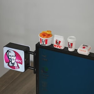 Puede incluir: Un letrero cuadrado negro de KFC con el logo del Coronel Sanders fijado al lado de un monitor de computadora. Sobre el marco del monitor hay cuatro artículos miniatura de KFC: un cubo de pollo frito, una bolsa de papel, un vaso y una caja.