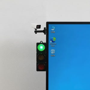 Puede incluir: Una cámara de seguridad blanca y un semáforo negro con una luz verde encendida están montados en el lateral de un monitor de ordenador. La pantalla del monitor muestra un fondo de escritorio azul con iconos para Este equipo, Microsoft Edge y Papelera.