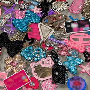 May include: A collection of assorted decorative charms and keychains featuring various shapes including bows, stars, flowers, and character faces. Many items are embellished with colorful rhinestones, glitter, and metallic finishes in pink, blue, and black.