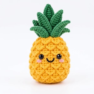 May include: A crocheted pineapple toy featuring a bright yellow body with a textured scale pattern and a green leafy top. The pineapple has a friendly face with two black circular eyes, a curved black stitched mouth, and small pink cheeks.