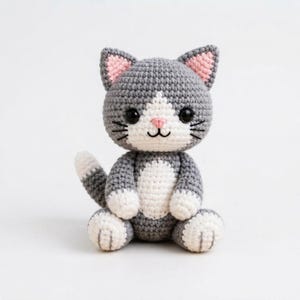Könnte beinhalten: Ein kleines, handgefertigtes Häkel-Katzenspielzeug aus grauem und weißem Garn. Die Katze hat eine weiße Brust, weiße Pfoten und eine weiße Schnauze mit einer kleinen rosa Nase. Sie besitzt große schwarze Sicherheitsaugen, spitze Ohren mit rosa Details und schwarze gestickte Schnurrhaare.