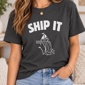 May include: A charcoal gray short-sleeve t-shirt featuring the white text SHIP IT above a graphic of a small sailboat with a coding symbol on its hull, depicted sailing over the edge of a waterfall into a pool of water below.