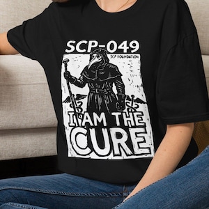 May include: A black short-sleeved t-shirt featuring a white graphic print of a plague doctor figure holding a staff. The text reads SCP-049, SCP FOUNDATION, and I AM THE CURE. The design includes two caduceus symbols on the sides of the figure.