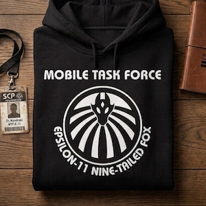 Op de afbeelding: Een zwarte hoodie met witte tekst die MOBILE TASK FORCE en EPSILON-11 NINE-TAILED FOX leest, gecentreerd rond een cirkelvormige afbeelding van een gestileerde vossenkop. Een zwart koord met een SCP Foundation ID-badge bevindt zich aan de linkerkant.