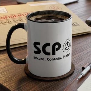May include: A white ceramic mug with a black handle and rim, featuring the black text SCP and Secure. Contain. Protect. along with a circular logo. The mug sits on a dark brown circular coaster on a wooden desk surface near a notebook and pen.