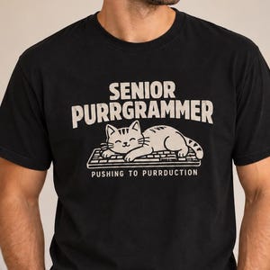 Könnte beinhalten: Ein schwarzes Kurzarm-T-Shirt mit einer Grafik einer Katze, die auf einer Computertastatur liegt. Der Text SENIOR PURRGRAMMER steht über der Katze und PUSHING TO PURRDUCTION unter der Tastatur in einer hellen Schrift im Used-Look.