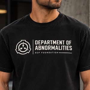 Op de afbeelding: Een zwart T-shirt met korte mouwen en een ronde hals, voorzien van een witte grafische print op de borst. Het ontwerp bevat het cirkelvormige logo van de SCP Foundation, gevolgd door de tekst DEPARTMENT OF ABNORMALITIES en SCP FOUNDATION.