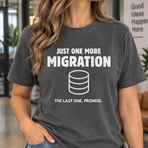 May include: A charcoal gray short-sleeve t-shirt featuring white text that reads JUST ONE MORE MIGRATION THE LAST ONE. PROMISE. above and below a white line-art graphic of a stacked database cylinder icon centered on the chest area.