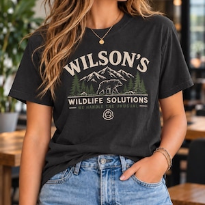 Peut inclure: Un t-shirt gris anthracite à manches courtes avec une impression graphique portant le texte WILSON'S WILDLIFE SOLUTIONS WE HANDLE THE UNUSUAL. Le motif comprend une chaîne de montagnes, des sapins, une silhouette d'ours et un emblème circulaire.