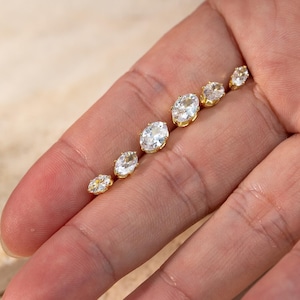May include: A row of six oval-shaped clear gemstones set in gold-toned metal prongs, arranged in a line from smallest to largest, showcasing faceted surfaces that reflect light with a brilliant, sparkling appearance.