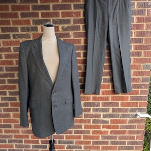 May include: A gray suit, consisting of a blazer and trousers, displayed on a mannequin and a hanger against a brick wall. The blazer has a two-button closure and two front pockets. The trousers are pleated and have a belt. The suit is made of a textured fabric.