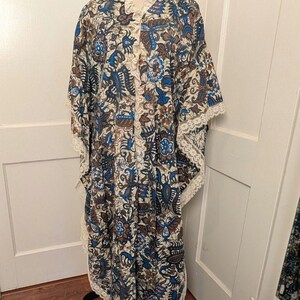 May include: A long, flowing robe with a floral pattern in shades of blue, brown, and cream. The robe features a lace trim along the collar, sleeves, and hem. The garment is displayed on a mannequin.