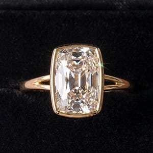 Puede incluir: Un anillo de oro con una gran piedra preciosa transparente de corte cojín rectangular en un engaste de bisel liso. La banda está hecha de oro amarillo pulido y presenta un diseño de vástago dividido que se separa cerca de la piedra central.