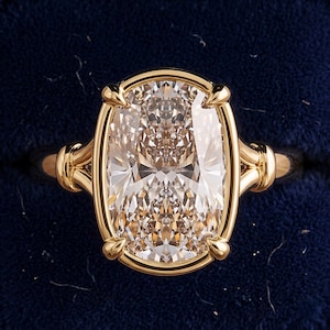 May include: A gold ring featuring a large, elongated cushion-cut champagne-colored gemstone set in a four-prong mounting. The band has a split-shank design that connects to the central setting, showcasing a polished metallic finish throughout.