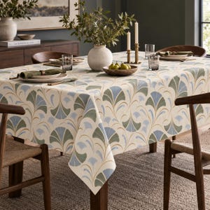 May include: A rectangular tablecloth featuring a repeating fan-shaped leaf pattern in muted shades of sage green, light blue, and gold on an off-white background. The fabric drapes over a dining table set with plates, glasses, and a bowl.