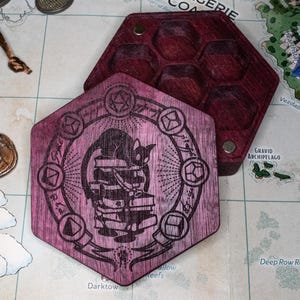 May include: A hexagonal purple wooden dice box with a lid featuring a laser-engraved illustration of a cat perched on a stack of books surrounded by a circular border of various polyhedral dice shapes and mystical symbols, with a six-compartment base.