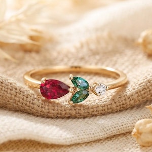 Puede incluir: Un anillo de oro con una piedra preciosa roja en forma de pera, dos piedras verdes de talla marquesa y una pequeña piedra clara redonda dispuestas en racimo sobre una banda fina. Las piedras forman un diseño delicado inspirado en la naturaleza.