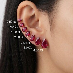 May include: A series of seven pear-shaped red gemstone stud earrings arranged in a row along the ear cartilage, labelled with sizes from 0.50 ct to 4.00 ct. Each stone is set in a gold-toned prong setting, showcasing a deep red colour and faceted cut.