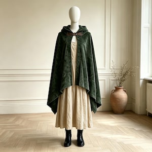 May include: A dark green velvet hooded cloak featuring a brown leather-like closure at the neck, worn over a beige midi-length dress and black ankle boots. The cloak has a wide, draped silhouette and a hood, providing a layered appearance.