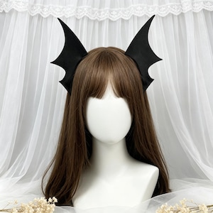 May include: A black headband featuring two upright bat wing attachments. The wings are pointed and have a smooth, matte finish. The headband is positioned on a mannequin head with long brown hair, showcasing the symmetrical wing design.