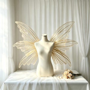 May include: A beige mannequin torso displays large, translucent fairy wings featuring intricate gold vein patterns and a subtle glitter finish. The wings are wide and pointed, extending outward from the shoulders of the form, which is set against a white backdrop.