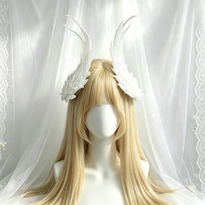May include: A white headpiece featuring two symmetrical, upward-curving wing-like structures with detailed feather textures. The accessory is positioned on a mannequin head with long, straight, light blonde hair and bangs, creating a striking contrast.