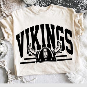 May include: A cream-coloured short-sleeve t-shirt featuring a bold black graphic print. The design displays the word VIKINGS in a large arched collegiate font above a detailed illustration of a horned helmet with horizontal lines below.