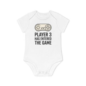 Op de afbeelding: Een witte babybody met korte mouwen voorzien van een afbeelding van een beige videogamecontroller boven de zwarte tekst PLAYER 3 HAS ENTERED THE GAME. Het kledingstuk heeft een ronde halslijn en drie drukknopen aan de onderkant als sluiting.