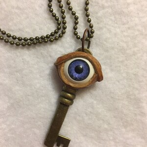 May include: A gold key necklace with a blue eye pendant. The eye is made of polymer clay and has a white iris and black pupil. The key is attached to a gold chain.