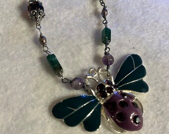 Bee pendant in enameled purple and green.  Wire wrapped necklace in green and purple natural stones.  Approximately 21”