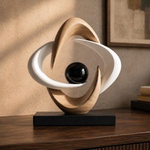 Può includere: Una scultura decorativa con una sfera nera lucida centrale sospesa all'interno di forme curve intrecciate. La struttura è composta da due archi in legno chiaro liscio e due archi bianchi opachi, montati su una base rettangolare nera.