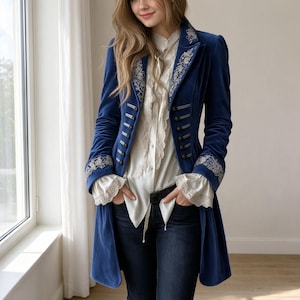 May include: A long-sleeved blue velvet coat featuring silver embroidery on the lapels and cuffs, paired with a cream-colored blouse with lace trim and dark blue jeans. The coat has a structured collar and multiple dark buttons along the front.