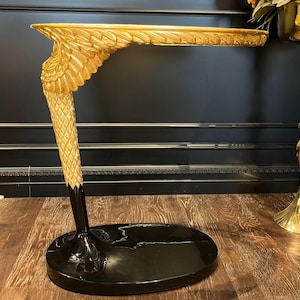 May include: A C-shaped side table featuring a gold-coloured base sculpted into the form of a feathered wing and leg. The table has a smooth, flat, circular top and rests on a solid, glossy black oval base that provides stability for the structure.
