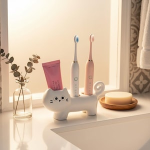 Puede incluir: Un soporte para cepillos de dientes de cerámica blanca con forma de gato y cara estilizada, que sostiene un tubo de pasta de dientes rosa, un cepillo de dientes eléctrico blanco y uno rosa claro. El soporte cuenta con una cola enroscada.