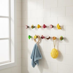 Peut inclure: Une collection de patères murales en forme de divers fruits et légumes, notamment un avocat, un citron, un fruit du dragon, une banane, une grenade et une noix de coco. Une serviette bleu clair est suspendue à un anneau en bois et une éponge jaune.