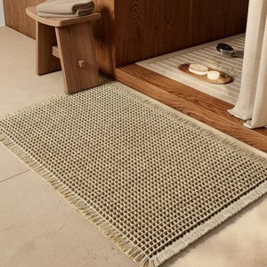 Op de afbeelding: Een rechthoekige beige badmat met een getextureerd wafelpatroon en franjes aan de randen. De mat is gemaakt van een geweven stof met een rasterachtig oppervlak, wat zorgt voor een tactiele afwerking. Het ontwerp bevat een rand met franjes.