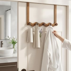 May include: A wooden over-the-door hook rack with a wavy design and five rounded pegs. The rack is mounted on a white door and holds two beige fabric towels and a white bathrobe. The wood has a natural brown finish with visible grain patterns.