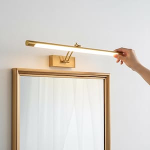 Könnte beinhalten: Eine goldfarbene Bilderleuchte aus Metall mit einem langen, horizontalen zylindrischen Lampenkopf, der helles weißes Licht ausstrahlt. Die Leuchte ist über einem rechteckigen Spiegel mit passendem Goldrahmen montiert und hat einen verstellbaren Arm.