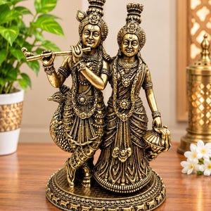 May include: A metallic gold-toned statue depicting two figures standing together on a circular base. One figure holds a flute, while the other stands beside them. Both are adorned with intricate jewellery, crowns, and traditional garments, with a peacock figure positioned behind the flute-playing figure.