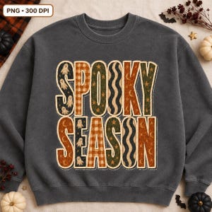 May include: A charcoal gray crewneck sweatshirt featuring the text SPOOKY SEASON in large block letters. The letters are filled with various patterns including ghosts, orange and white plaid, green stars, and wavy lines in orange and black.