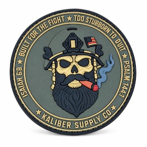Könnte beinhalten: Ein kreisförmiger olivgrüner Aufnäher mit einem goldenen Totenkopf mit schwarzem Bart und Schnurrbart, der einen taktischen Helm mit einer kleinen amerikanischen Flagge trägt. Eine Zigarre mit blauem Rauch ist im Mund. Text: BUILT FOR THE FIGHT, TOO STUBBORN TO QUIT, ISAIAH 6:8, PSALM 144:1, KALIBER SUPPLY CO.