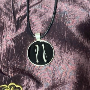 May include: A circular silver-toned pendant featuring two small, white, elongated bone-like shapes set against a smooth black background. The pendant is attached to a thin, black cord necklace that extends upward from the top metal bail.