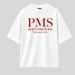 Può includere: Una t-shirt bianca a maniche corte con scollo rotondo. La parte anteriore presenta una scritta rosso scuro che recita PMS, seguita dalla frase don't touch me, e dal testo più piccolo if you want to live. Il capo ha una vestibilità ampia e un'etichetta.