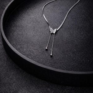Può includere: Una collana color argento con una catena a serpente piatta che si collega a un ciondolo a forma di farfalla. Due catenine delicate pendono dalla base della farfalla, terminando ciascuna con una piccola perlina argentata rotonda.