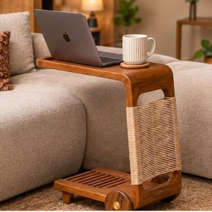 Puede incluir: Una mesa auxiliar de madera en forma de C con acabado liso, que presenta un panel vertical envuelto en cuerda trenzada de color claro, un estante inferior con listones y ruedas pequeñas. Un portátil plateado y una taza de cerámica blanca acanalada descansan sobre la superficie.
