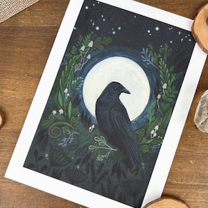 Peut inclure: Une impression d'art encadr&eacute;e repr&eacute;sentant un corbeau noir devant une pleine lune. Le corbeau est entour&eacute; d'un feuillage vert fonc&eacute;, de champignons blancs et d'&eacute;toiles sur un fond bleu fonc&eacute;. L'&oelig;uvre est sur fond blanc.