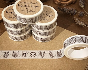 May include: A stack of six rolls of washi tape featuring a woodland animal theme. The tape is white with black and grey illustrations of animals such as owls, foxes, and badgers. The tape is labelled "Woodland Friends Washi Tape 10m alice darkling".