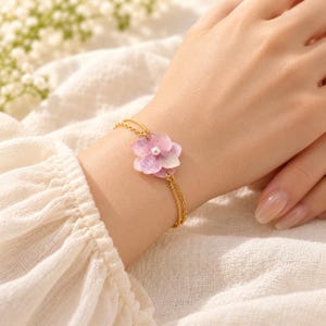 May include: A gold-toned double chain bracelet featuring a delicate pink and white hydrangea flower charm with a small white pearl at its center. The bracelet is secured with a thin metal chain that wraps around the wrist.