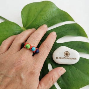 Op de afbeelding: Een ring met vier kleine, ronde, veelkleurige kralen in zwart, groen, turquoise en donkerblauw, vastgezet met een rood geknoopt koord. Daarnaast ligt een gladde witte steen met de tekst EL RINCÓN DE ANROS in bruin bedrukt.
