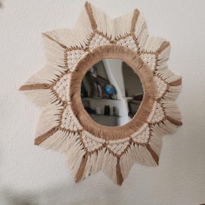 May include: A circular wall mirror framed by intricate macrame cord in cream and tan colours. The fibre art border features a starburst pattern with knotted details and long, fringed edges radiating outward from the central reflective glass surface.